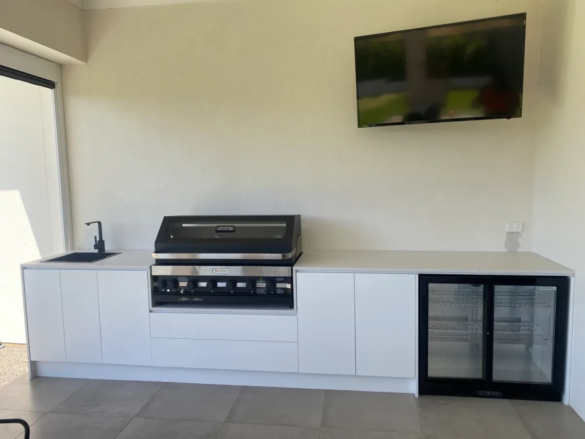 White outdoor kitchen with built-in BBQ, sink, drinks fridge and wall-mounted TV