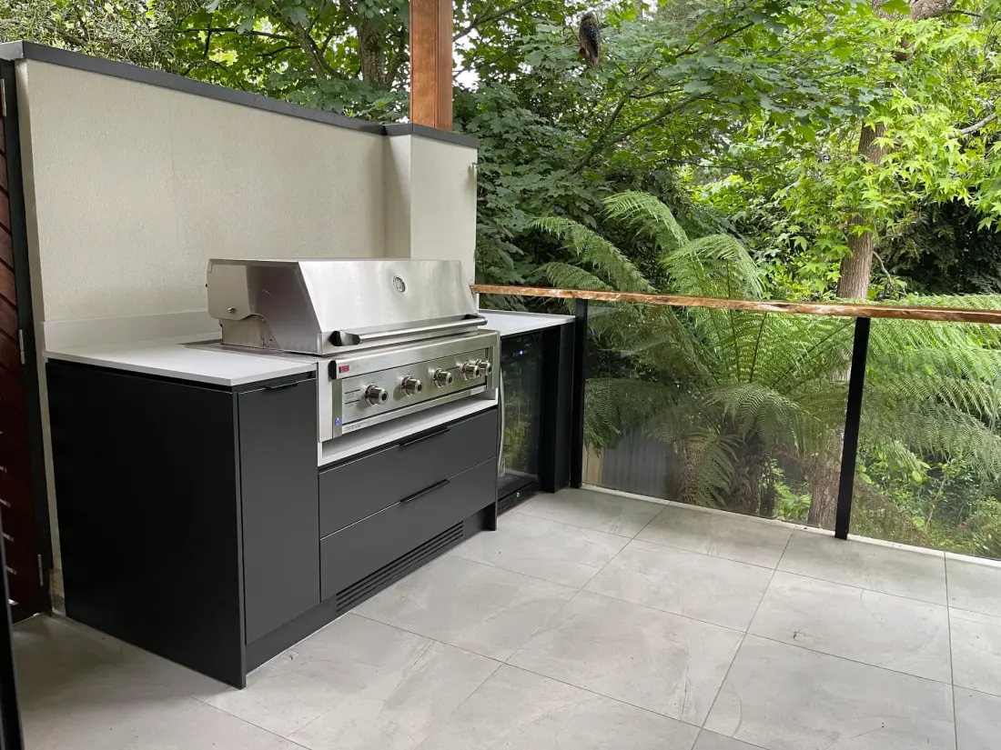 Dark outdoor kitchen with stainless steel BBQ, integrated fridge and garden backdrop